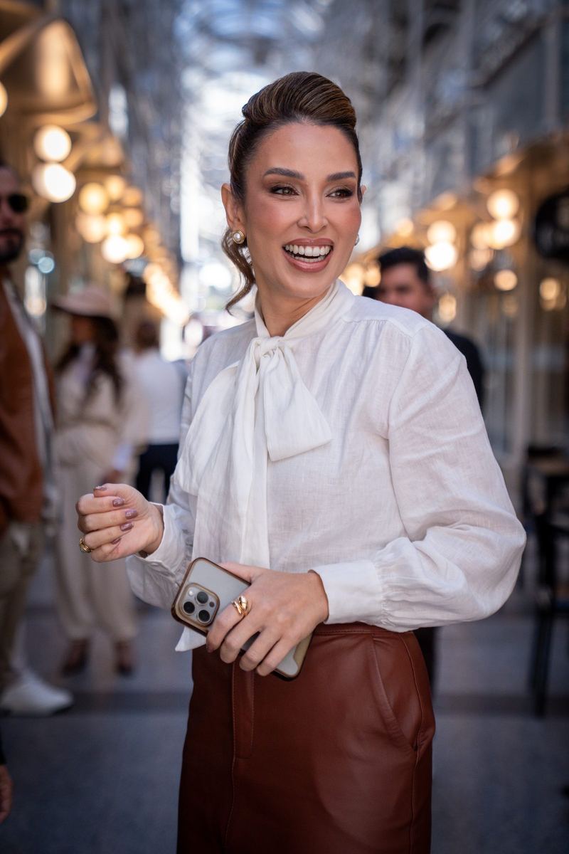 Mulher sorrindo, de blusa branca com laço, em galeria iluminada
