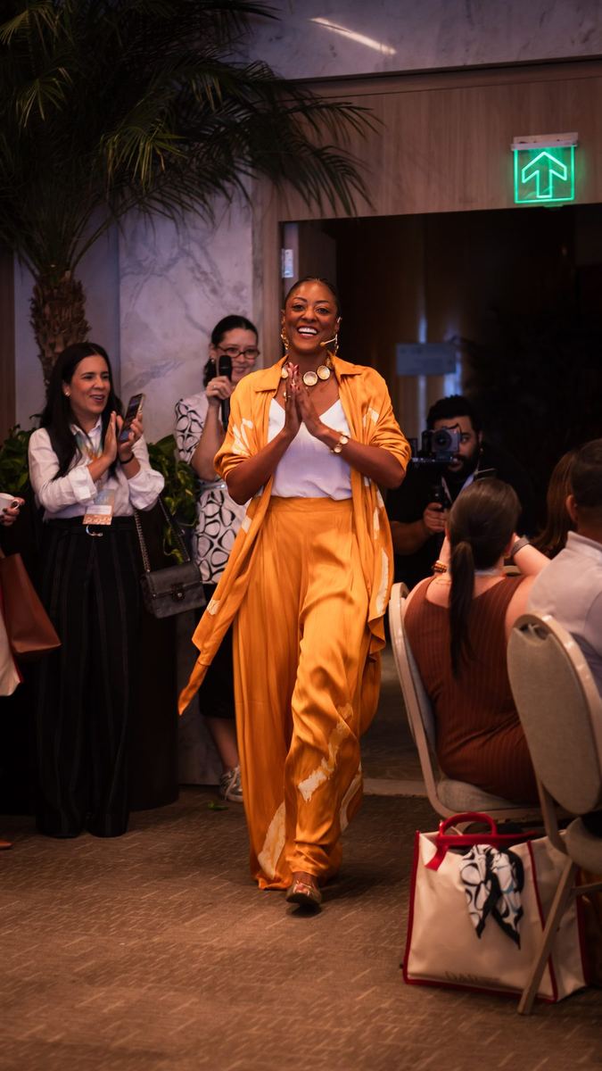 Palestrante de conjunto laranja entrando aplaudida em sala de evento