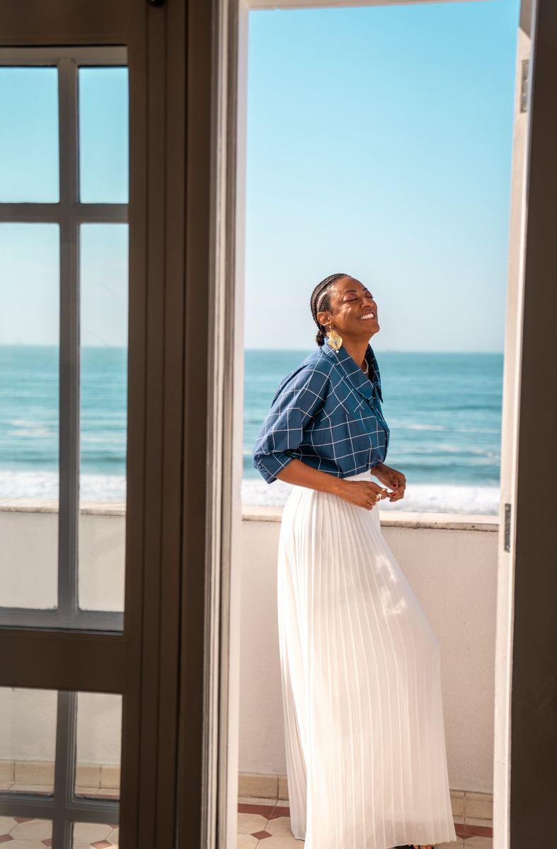 Mulher de camisa xadrez azul e saia plissada branca, sorrindo em janela com o mar ao fundo